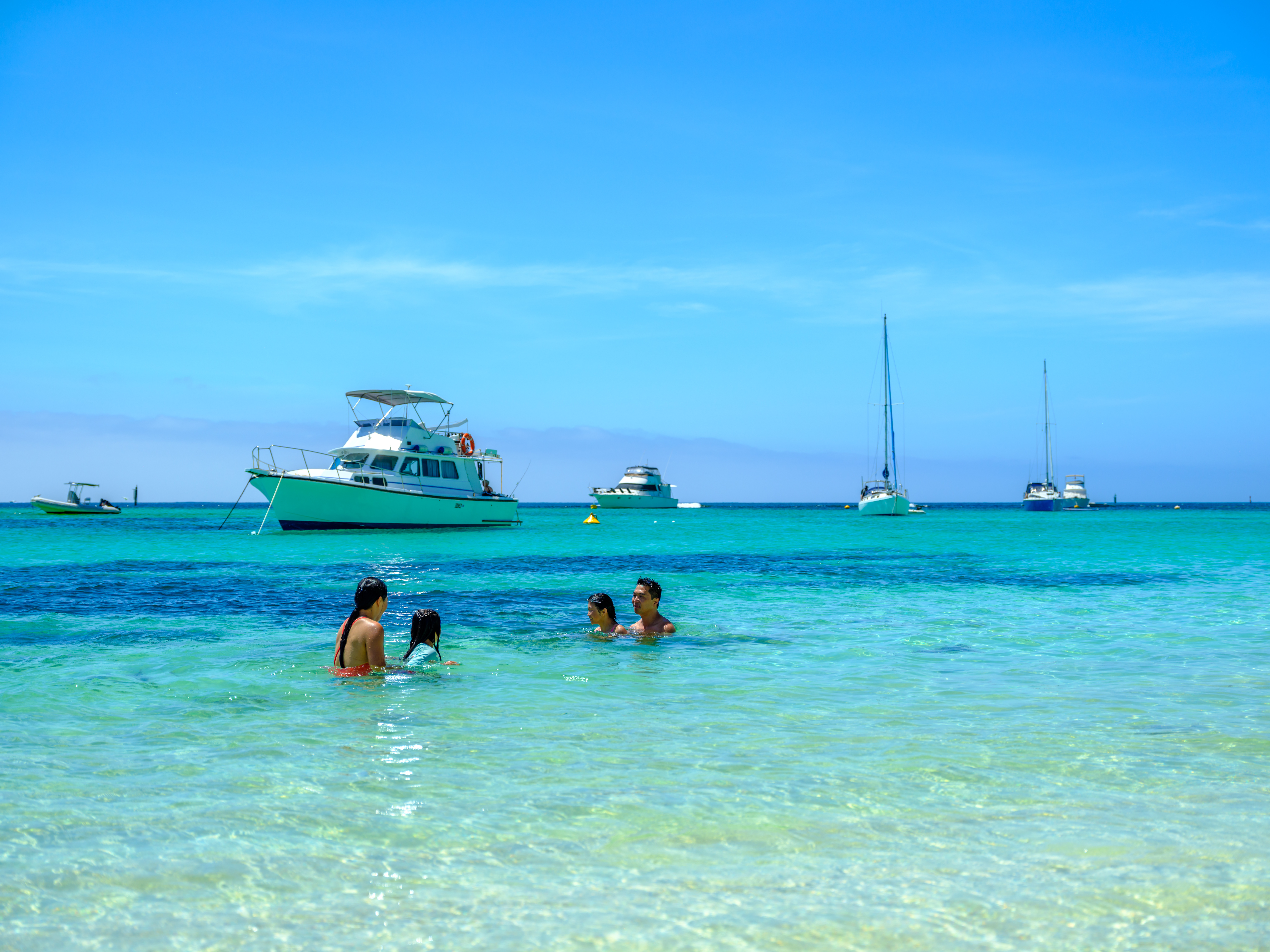 A family enjoys swimming in clear turquoise waters with boats anchored in the distance under a bright blue sky.